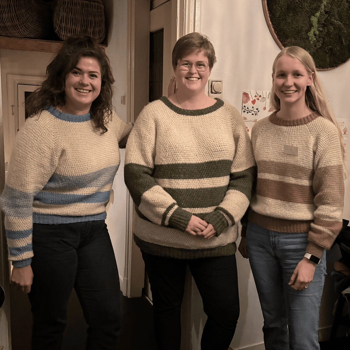 Drie vrouwen dragen zelfgemaakte gestreepte truien in blauw, groen en beige, binnen.