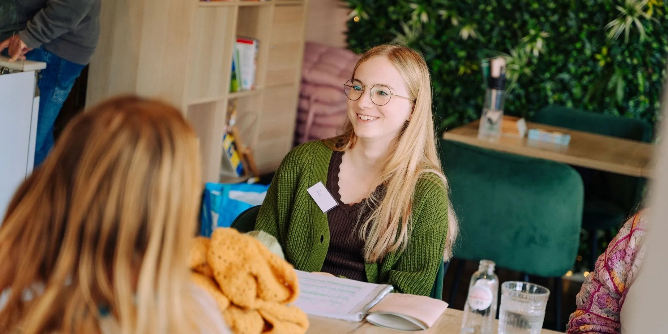 Lachende vrouw met bril en groen vest zit aan tafel tegenover iemand met geel haakwerk.