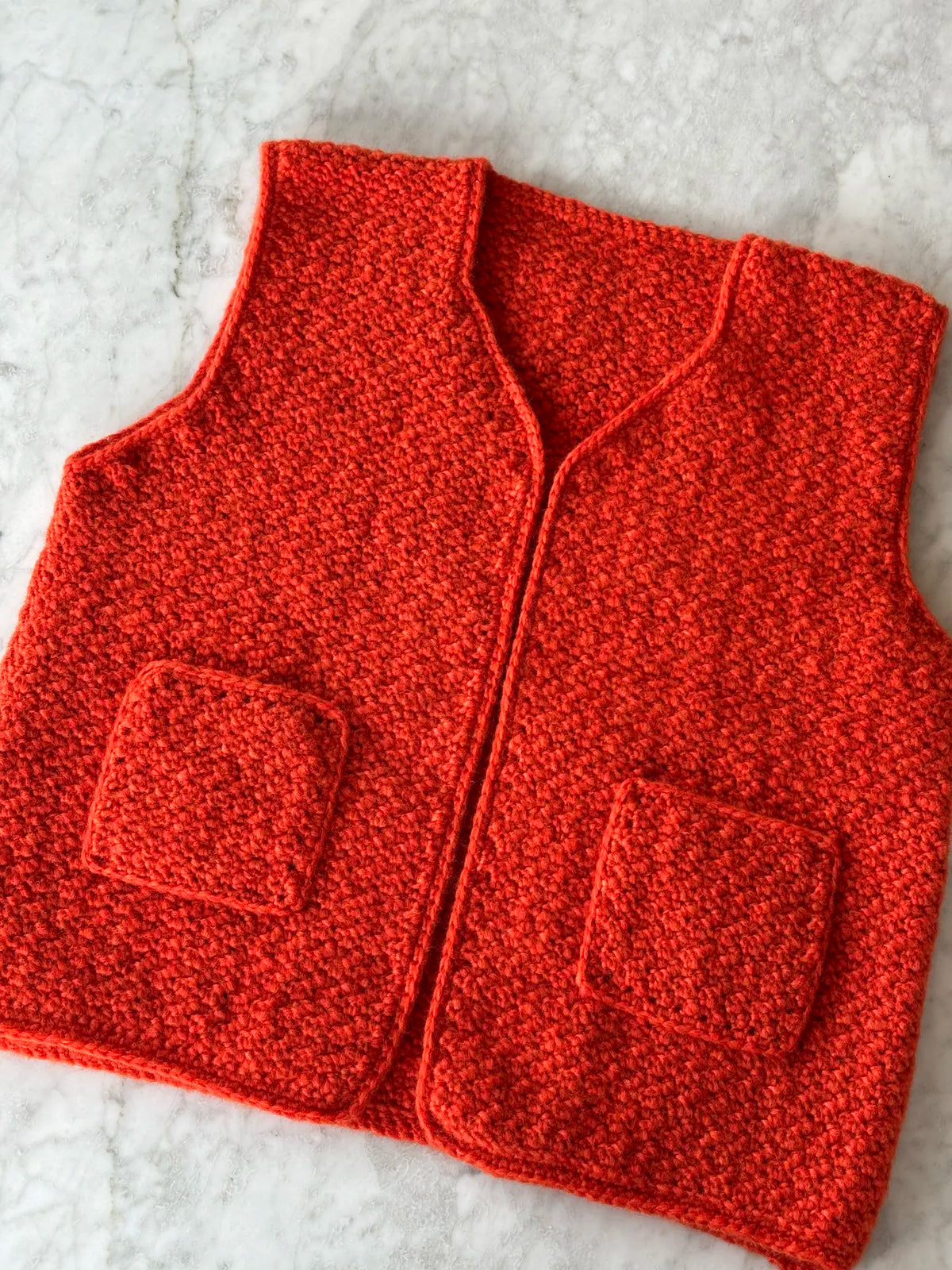 Gehaakte Nora bodywarmer in oranjerood met V-hals en twee opgestikte zakken op marmer