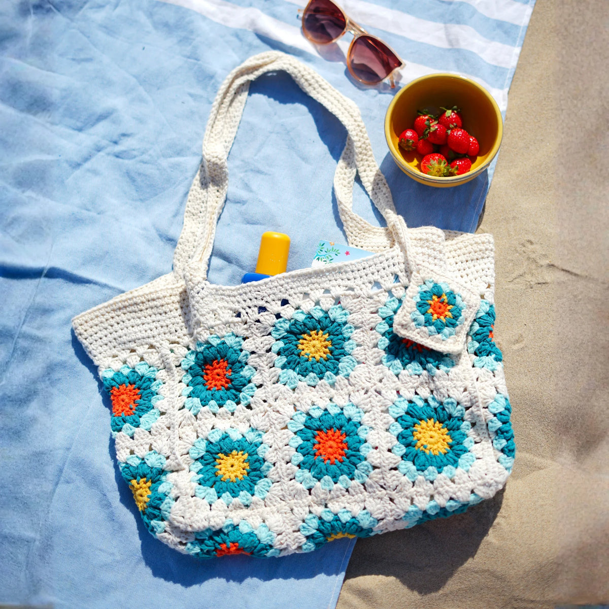 Gehaakte Sunny Beach Bag met granny squares in blauw, oranje en geel op strandlaken met zonnebril en aardbeien