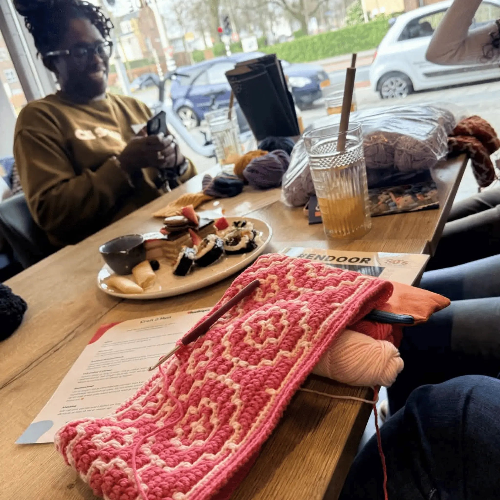 Roze-wit gehaakt werk op cafétafel, met garen, snacks en lachende vrouw op de achtergrond.