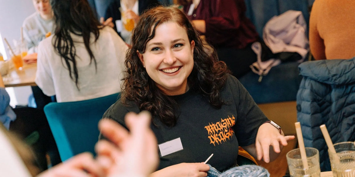 Lachende vrouw met donkere krullen haakt aan blauw garen tijdens een drukke workshop.