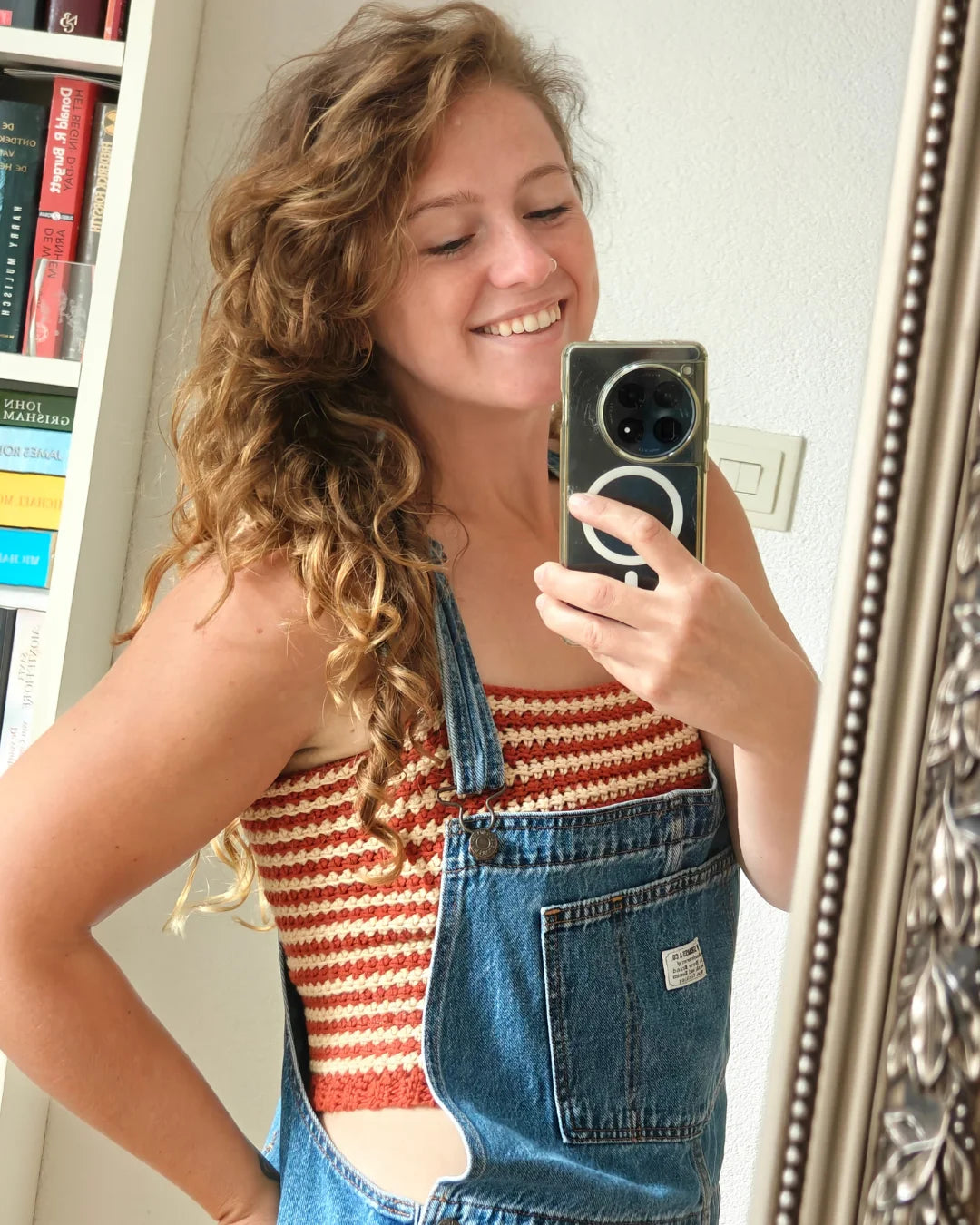 Lachende vrouw maakt spiegelselfie in gehaakte Striped Top met rood-beige strepen onder denim tuinbroek
