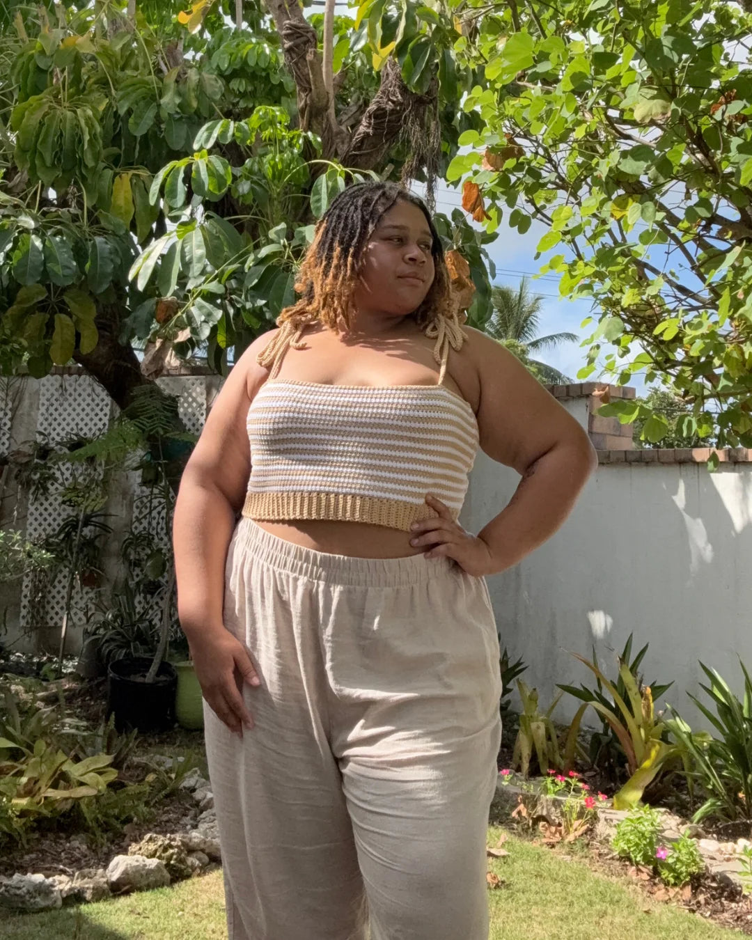 Vrouw draagt gehaakte Striped Top in oker en wit met beige broek in zonnige tuin