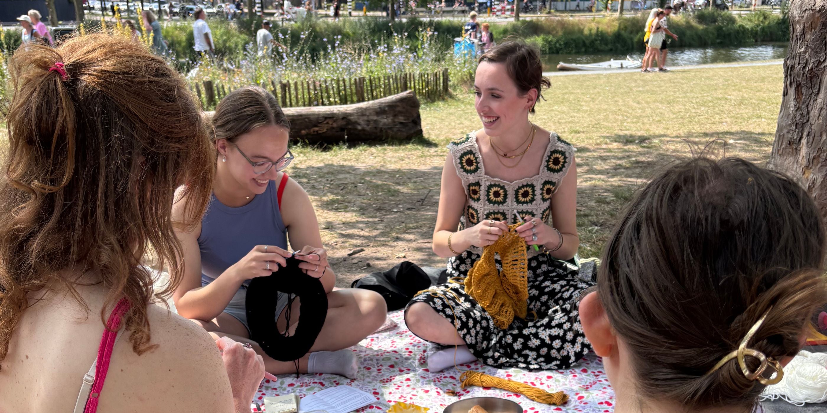Vier vrouwen haken samen op een picknickkleed in het gras bij het water.